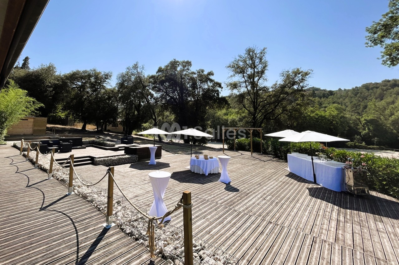Terrasse en bois aménagée avec tables hautes, parasols et mobilier, entourée de végétation sous un ciel dégagé.