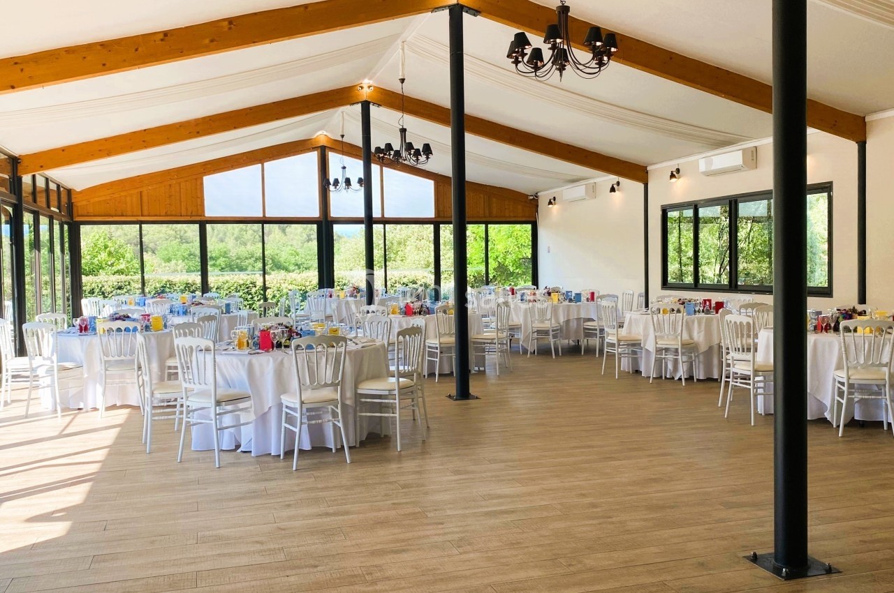 Salle lumineuse avec de grandes baies vitrées, tables rondes dressées avec nappes blanches et chaises alignées.