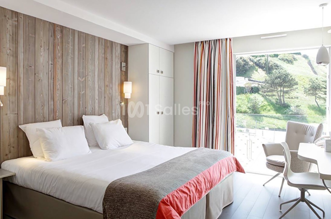 Chambre lumineuse avec lit double, mur en bois, rideaux colorés et vue sur un paysage verdoyant à travers une baie vitrée.