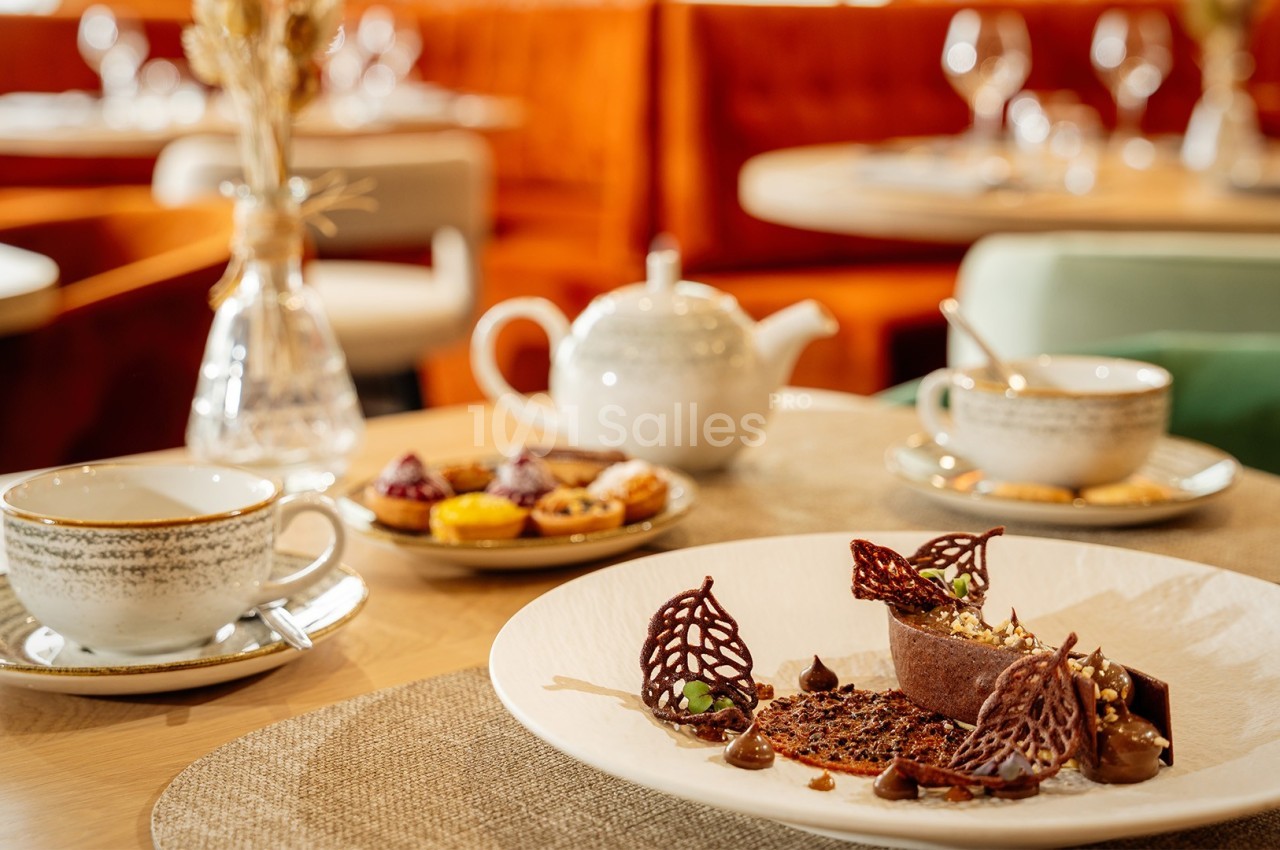 Dessert chocolaté décoré sur une assiette, accompagné de tasses de thé et de pâtisseries dans un restaurant élégant.