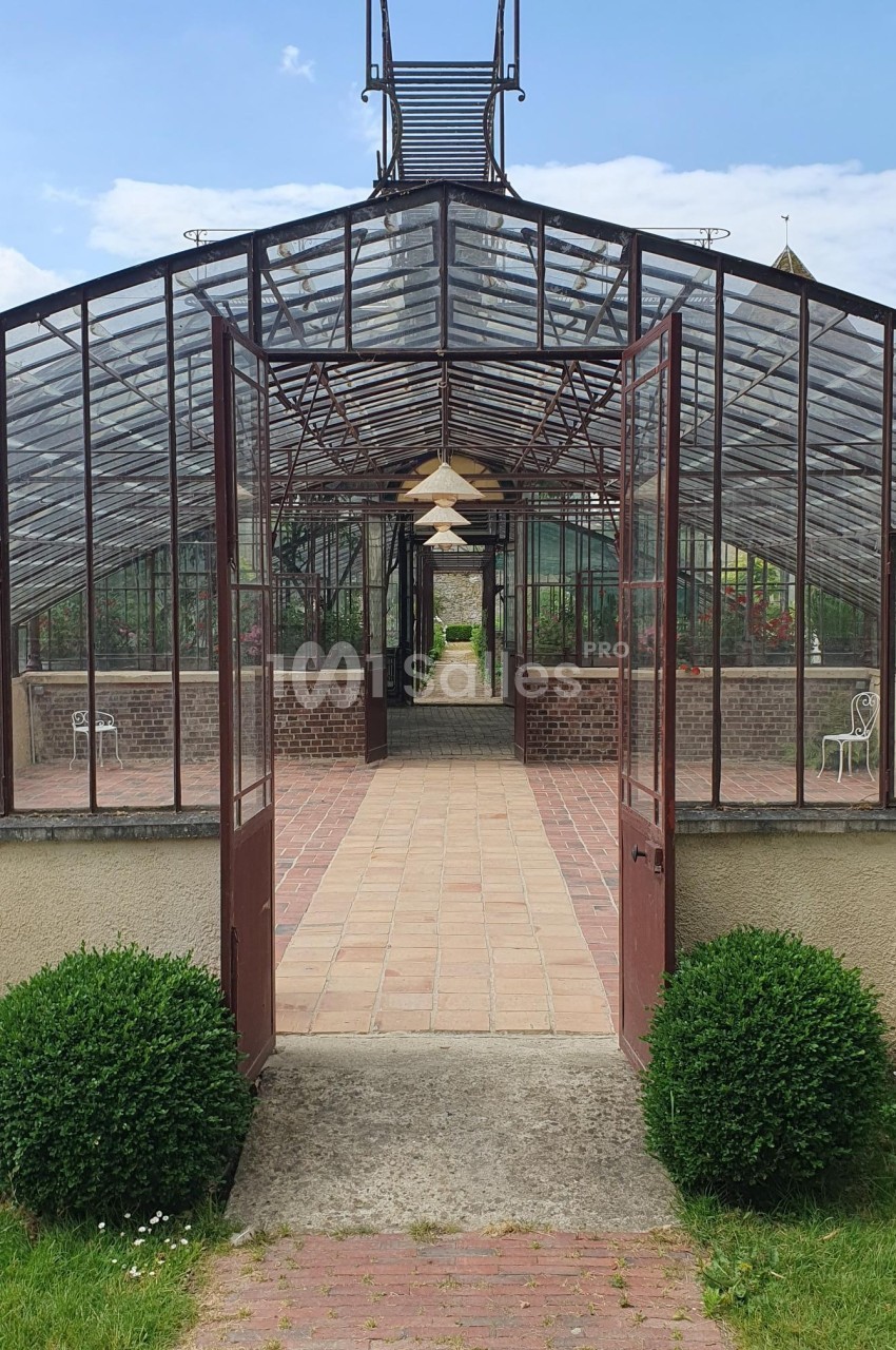Serre ancienne avec structure métallique et portes ouvertes, laissant voir un chemin pavé et des plantes à l'intérieur.