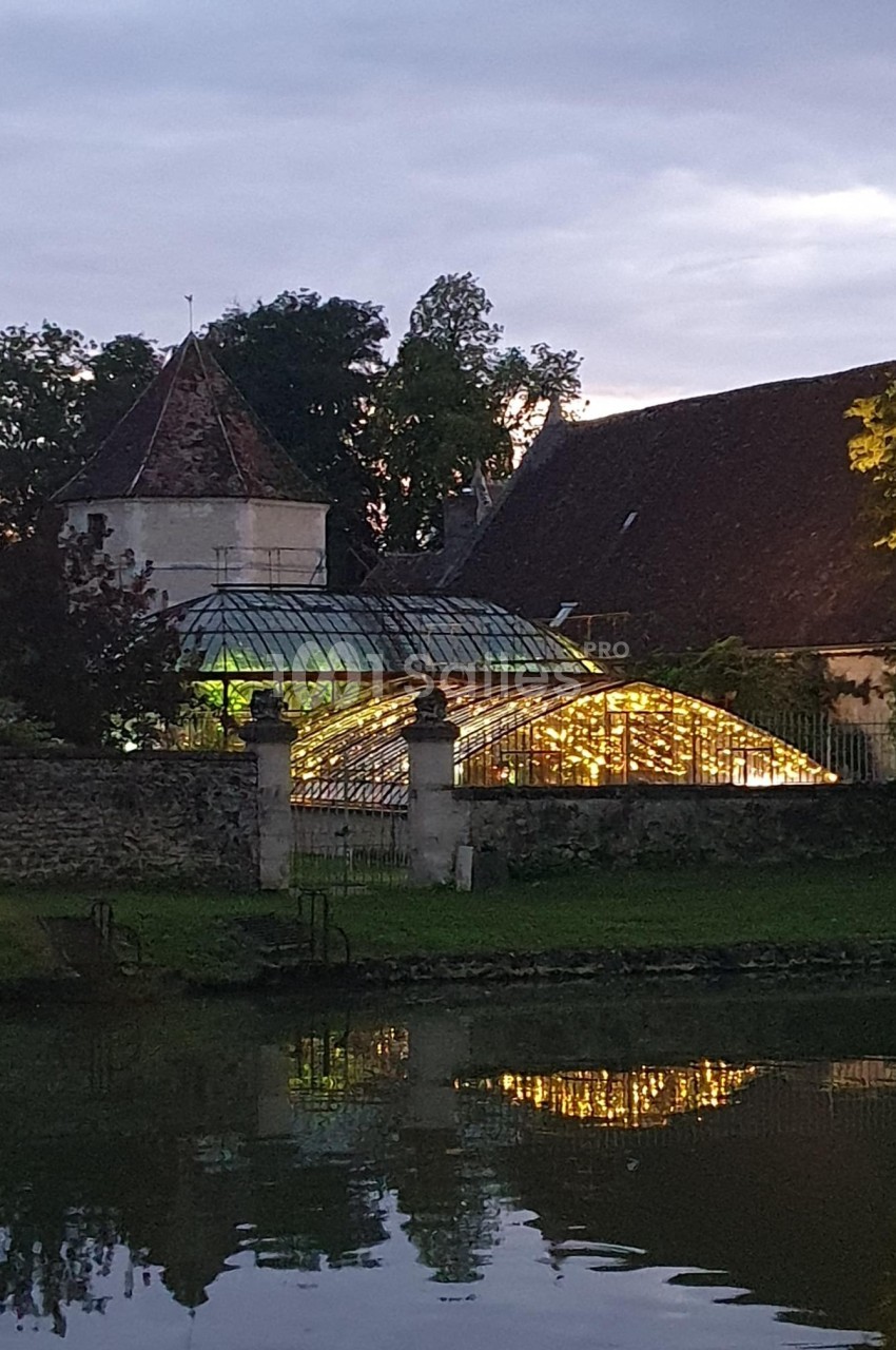 Serre éclairée au crépuscule, entourée de bâtiments anciens et d'arbres, avec un étang au premier plan.