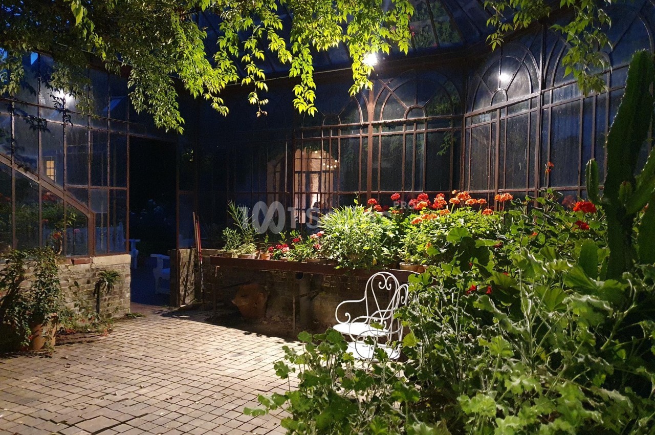 Serre ancienne éclairée le soir, avec des plantes luxuriantes, des fleurs colorées et une chaise blanche en fer forgé.
