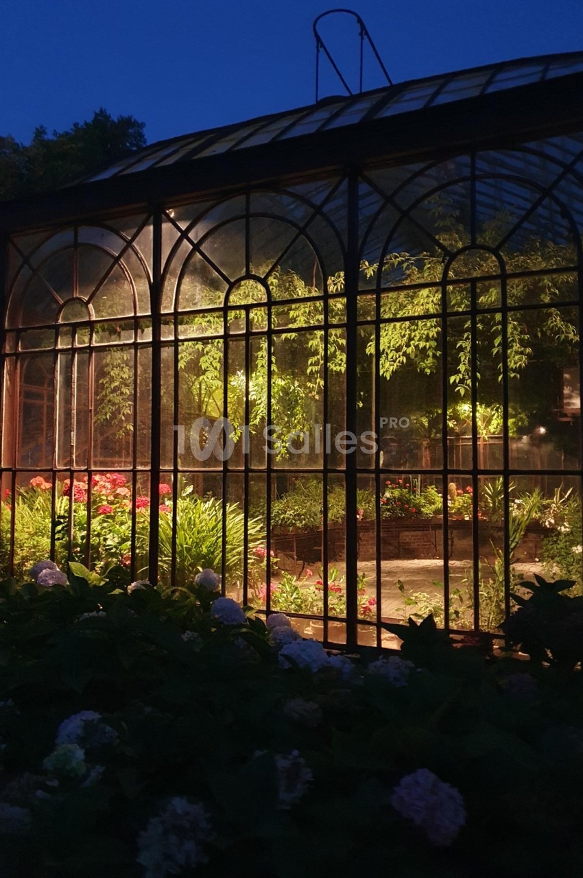 Serre éclairée de nuit, entourée de végétation, avec des plantes et fleurs visibles à l'intérieur.