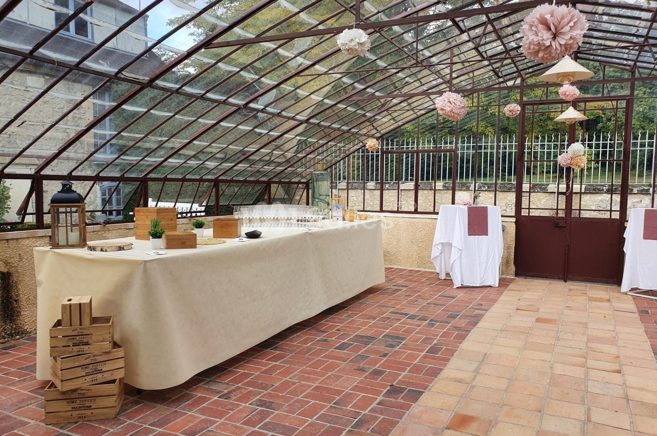 Salle vitrée avec une table de buffet décorée, des lanternes et des fleurs suspendues, sol en carreaux rouges et beiges.