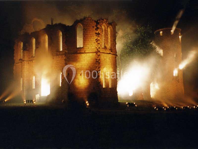 Ruines éclairées par des lumières chaudes dans un décor nocturne, avec de la fumée entourant les structures.