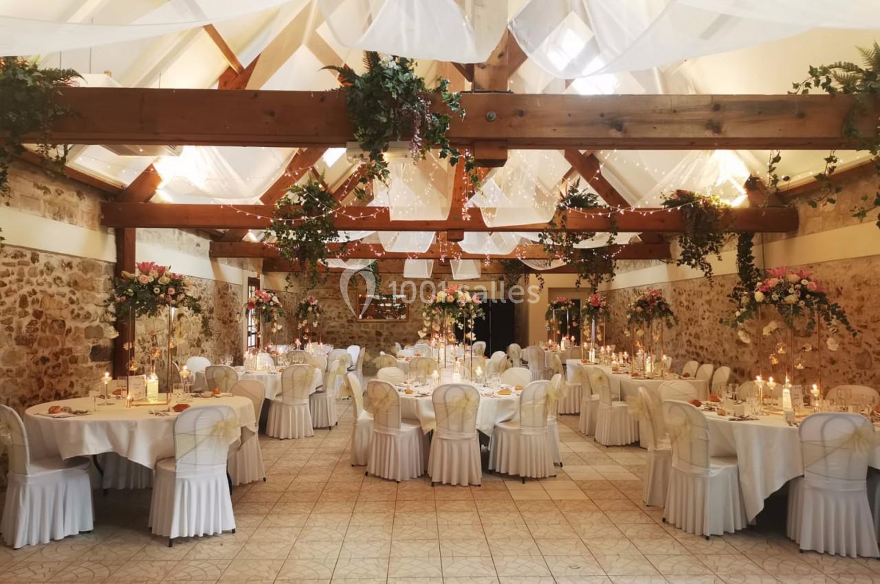 Salle de réception décorée avec des tables rondes, nappes blanches, chandeliers et guirlandes florales suspendues.