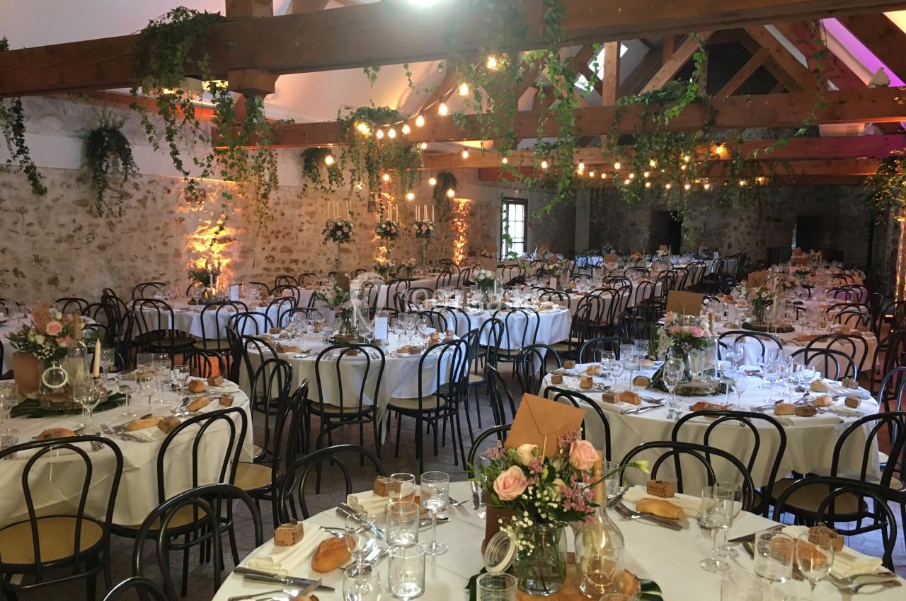 Salle de réception décorée avec des tables rondes dressées, des fleurs, des guirlandes lumineuses et des poutres apparentes.