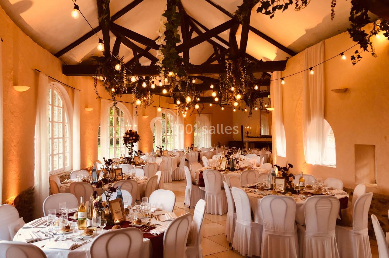 Salle de réception décorée avec des guirlandes lumineuses, des tables dressées et des chaises blanches.