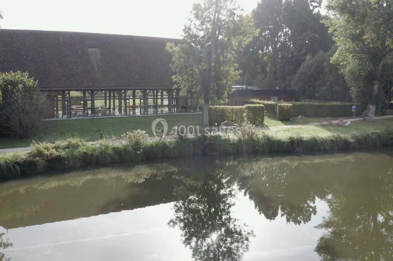 Vue d'un bâtiment à colombages entouré de verdure, avec un étang en premier plan reflétant le paysage.