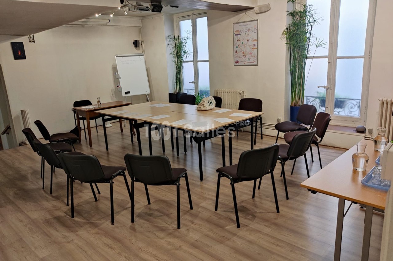 Salle de réunion lumineuse avec tables disposées en U, chaises autour, tableau blanc et plantes en arrière-plan.