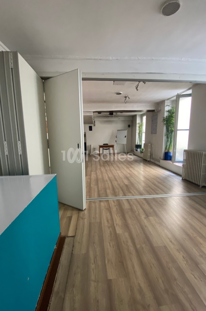 Salle lumineuse avec parquet, cloison ouverte, mobilier minimaliste et plantes près des fenêtres.