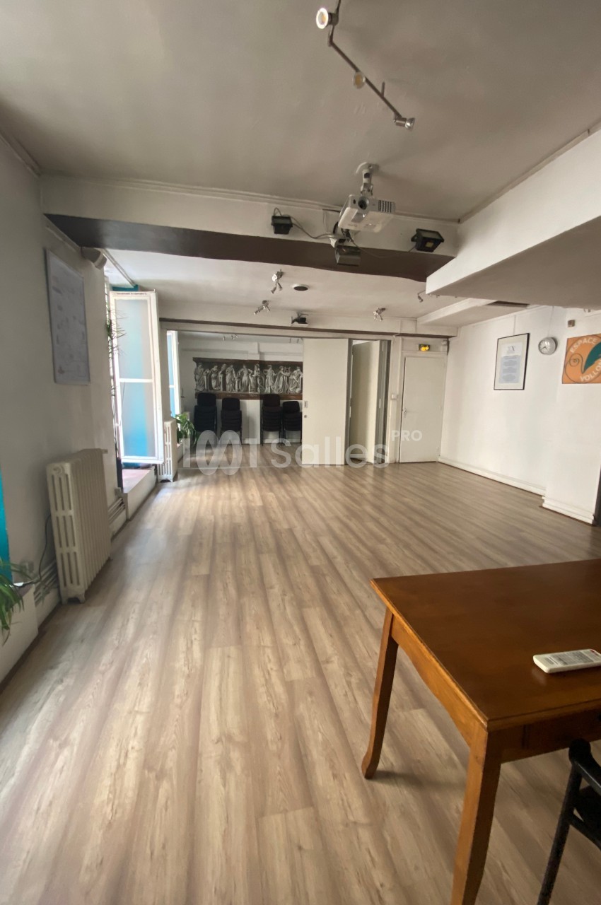 Salle lumineuse avec parquet clair, équipée d’un projecteur au plafond, des plantes et une table en bois au premier plan.