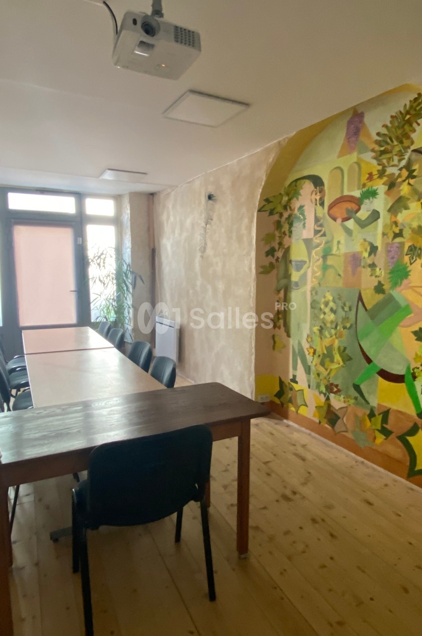 Salle de réunion lumineuse avec une grande table en bois, des chaises noires et une fresque colorée sur le mur.