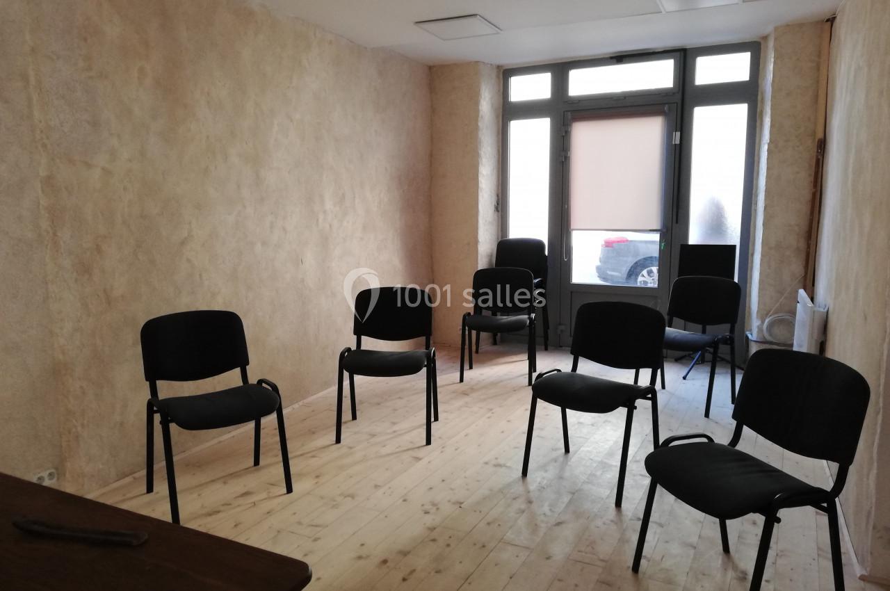 Salle lumineuse avec murs clairs, parquet en bois et plusieurs chaises noires disposées en cercle.