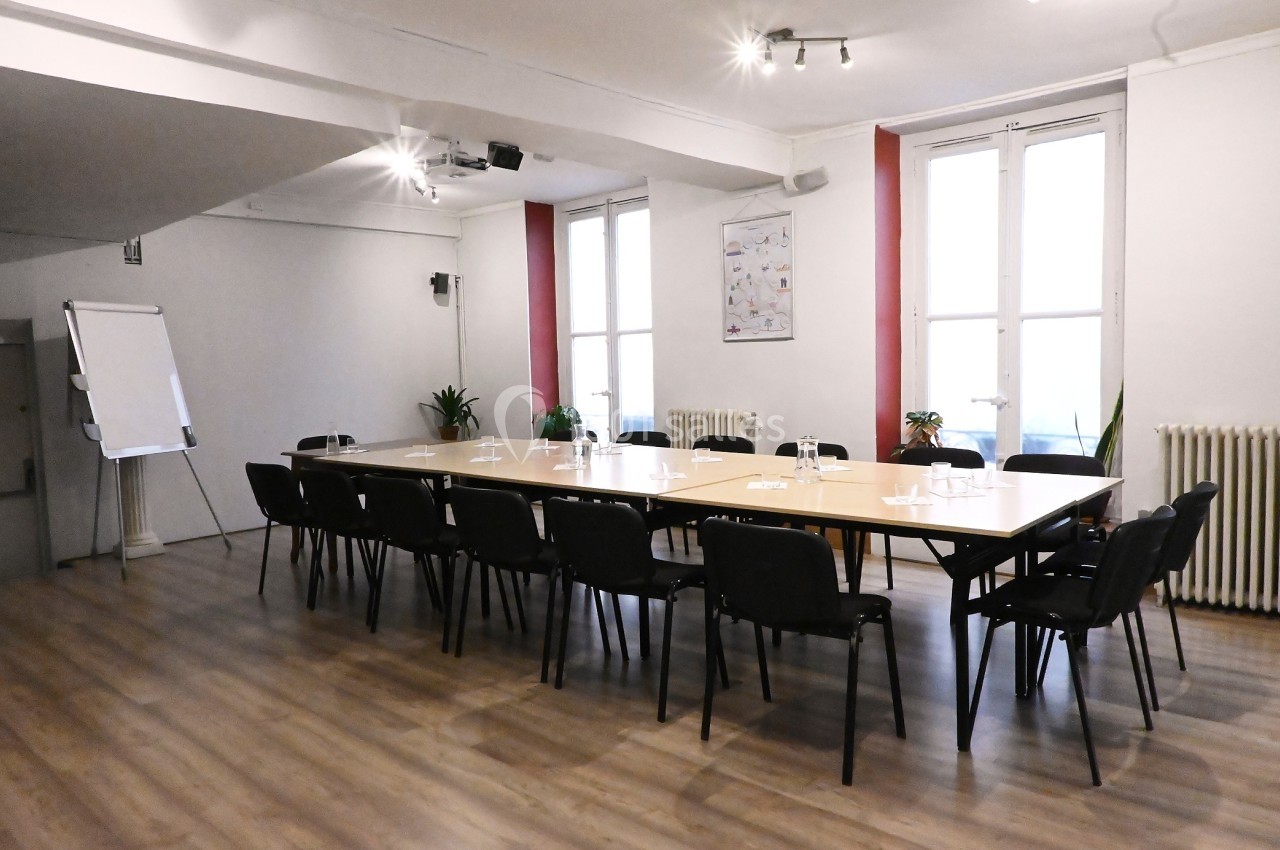 Salle de réunion lumineuse avec une grande table rectangulaire, chaises noires, tableau blanc et fenêtres.
