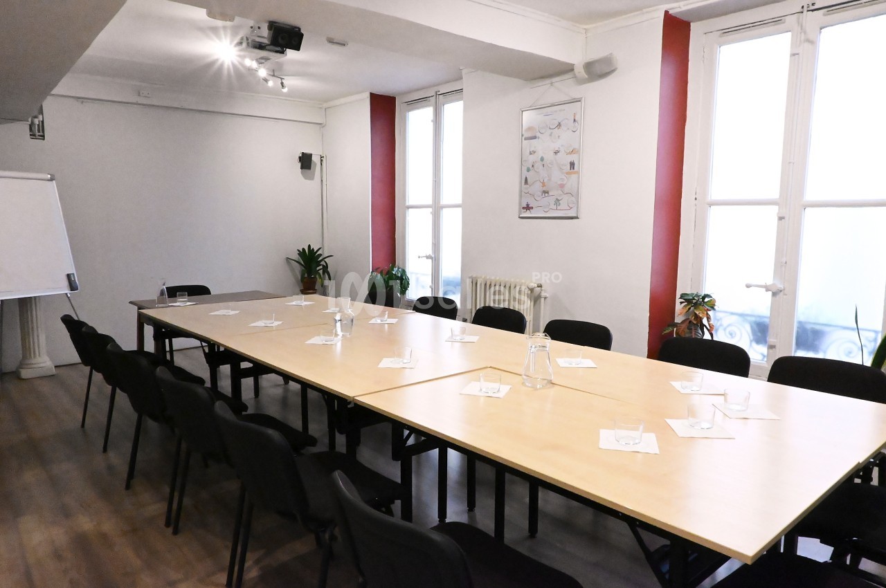 Salle de réunion lumineuse avec une grande table rectangulaire, chaises noires et quelques plantes près des fenêtres.
