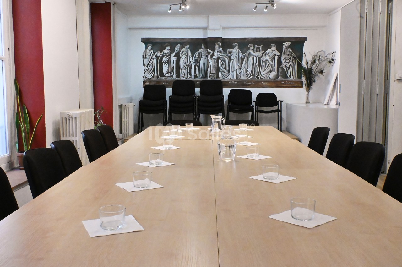 Salle de réunion avec une grande table en bois, chaises noires, verres disposés et fresque murale en arrière-plan.