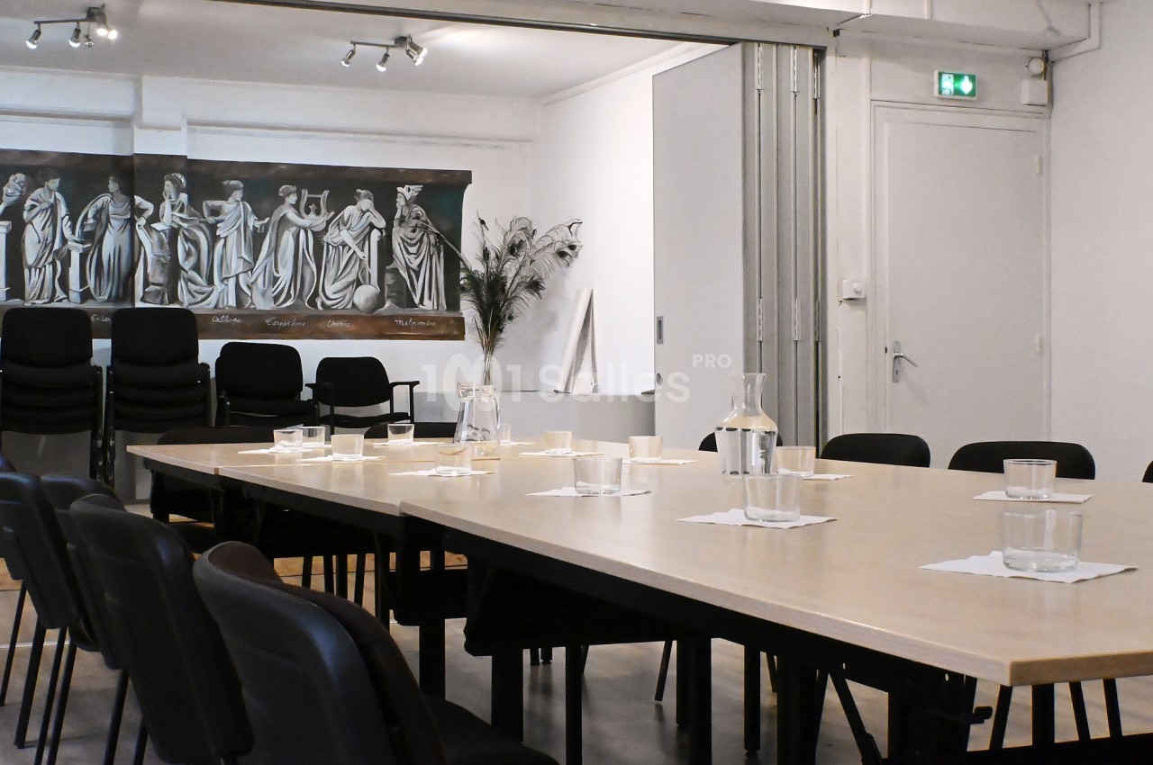 Salle de réunion avec une grande table, des chaises noires, des carafes d'eau et un tableau représentant des figures…