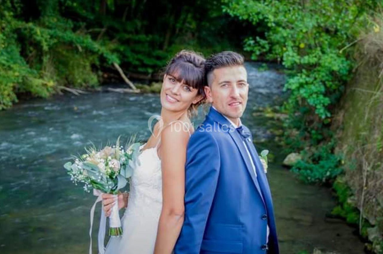 Un couple en tenue de mariage pose dos à dos près d'une rivière entourée de végétation.