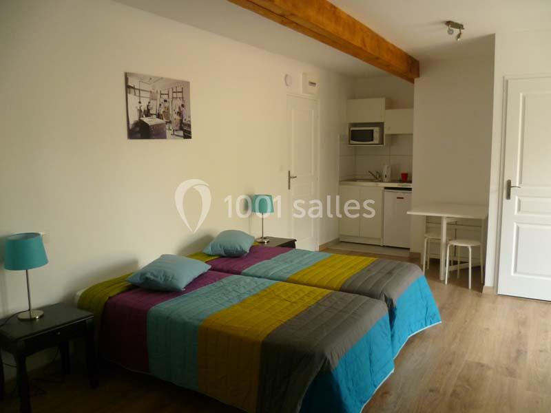 Chambre lumineuse avec lit double, kitchenette équipée, table et décoration simple sur parquet clair.