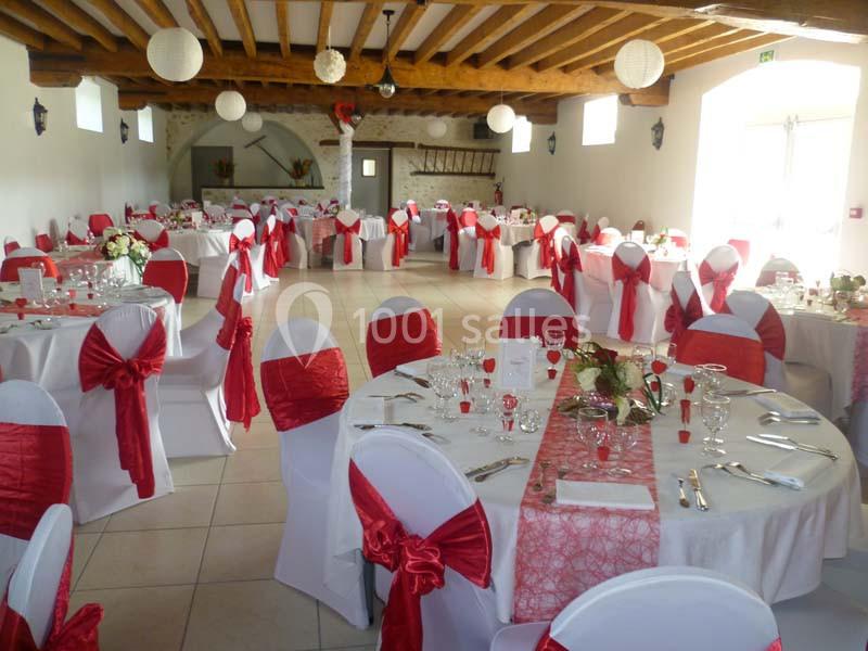 Salle de réception décorée avec des tables rondes, nappes blanches et rouges, chaises assorties et éclairage naturel.
