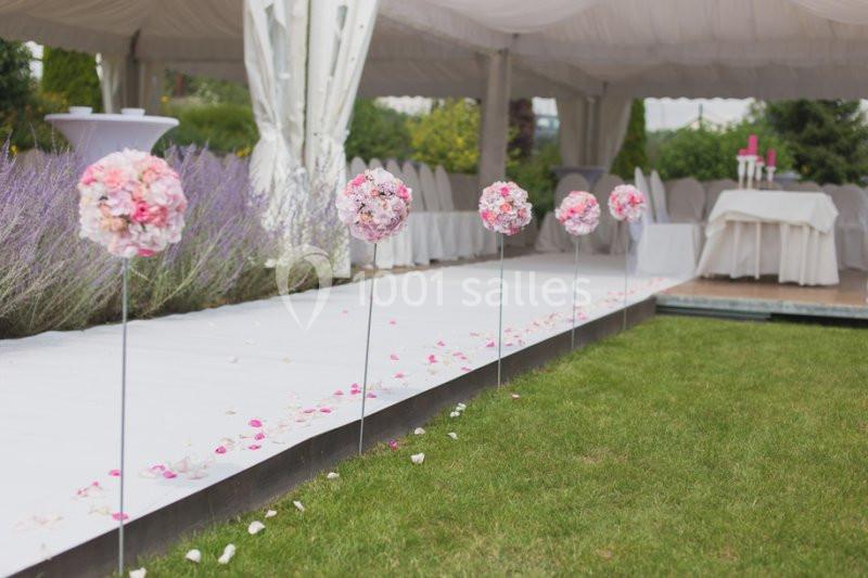 Allée décorée de boules de fleurs roses menant à une table sous une tente blanche, entourée de verdure.