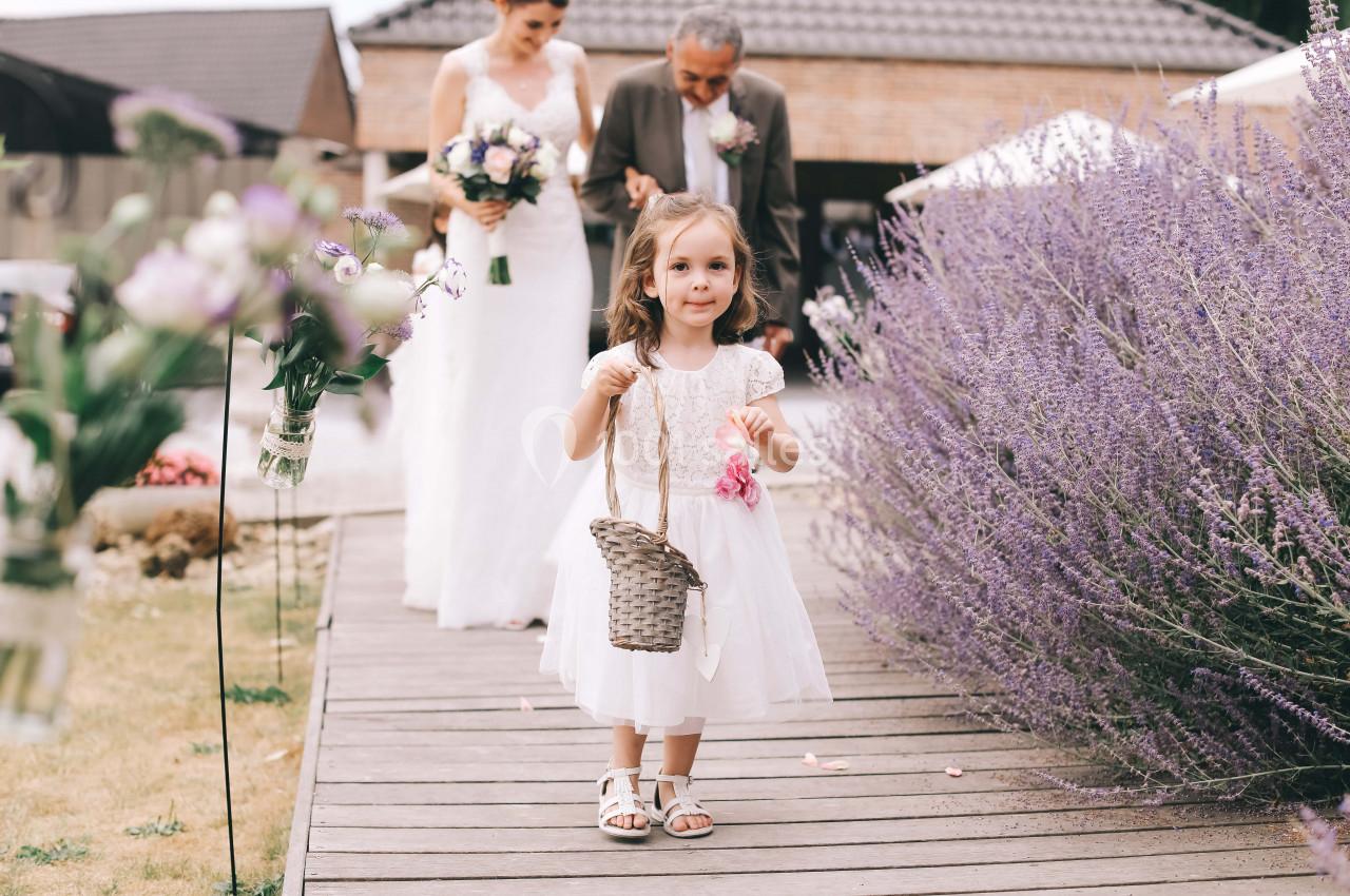 Une fillette en robe blanche marche sur un pont en bois, tenant un panier, suivie par une mariée et un homme.