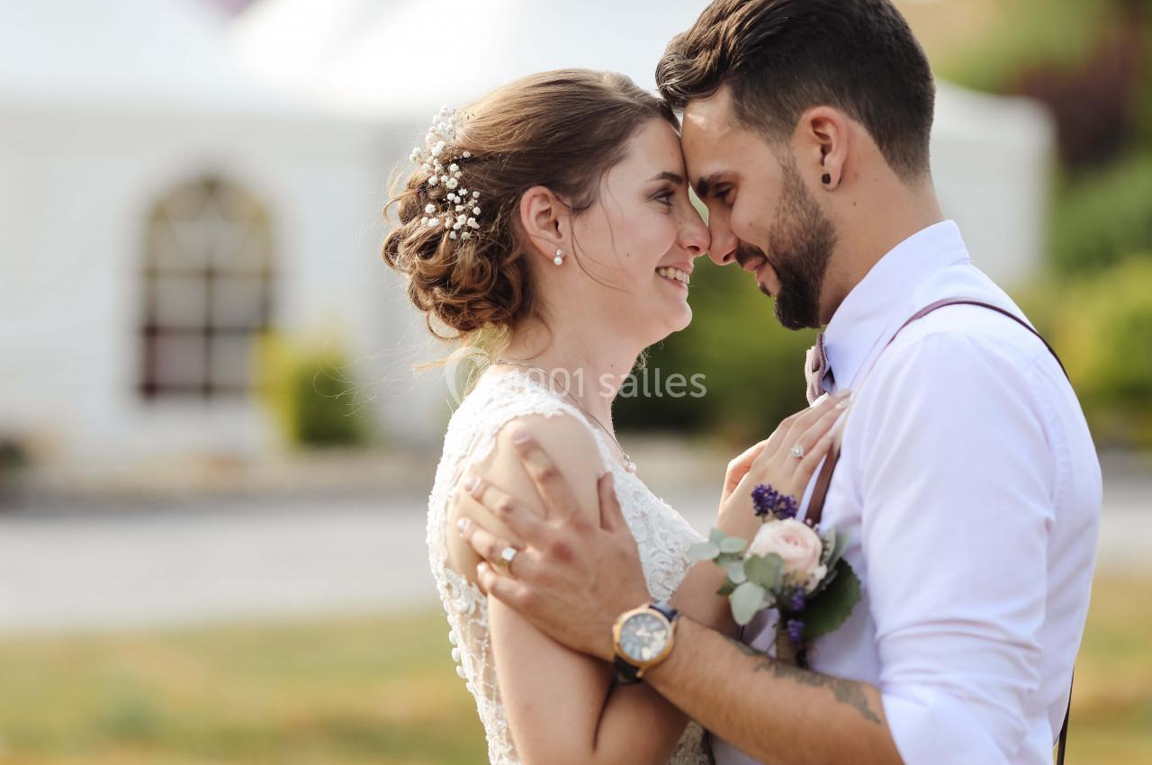 Un couple de jeunes mariés s'enlace tendrement en extérieur, devant une tente blanche floue en arrière-plan.