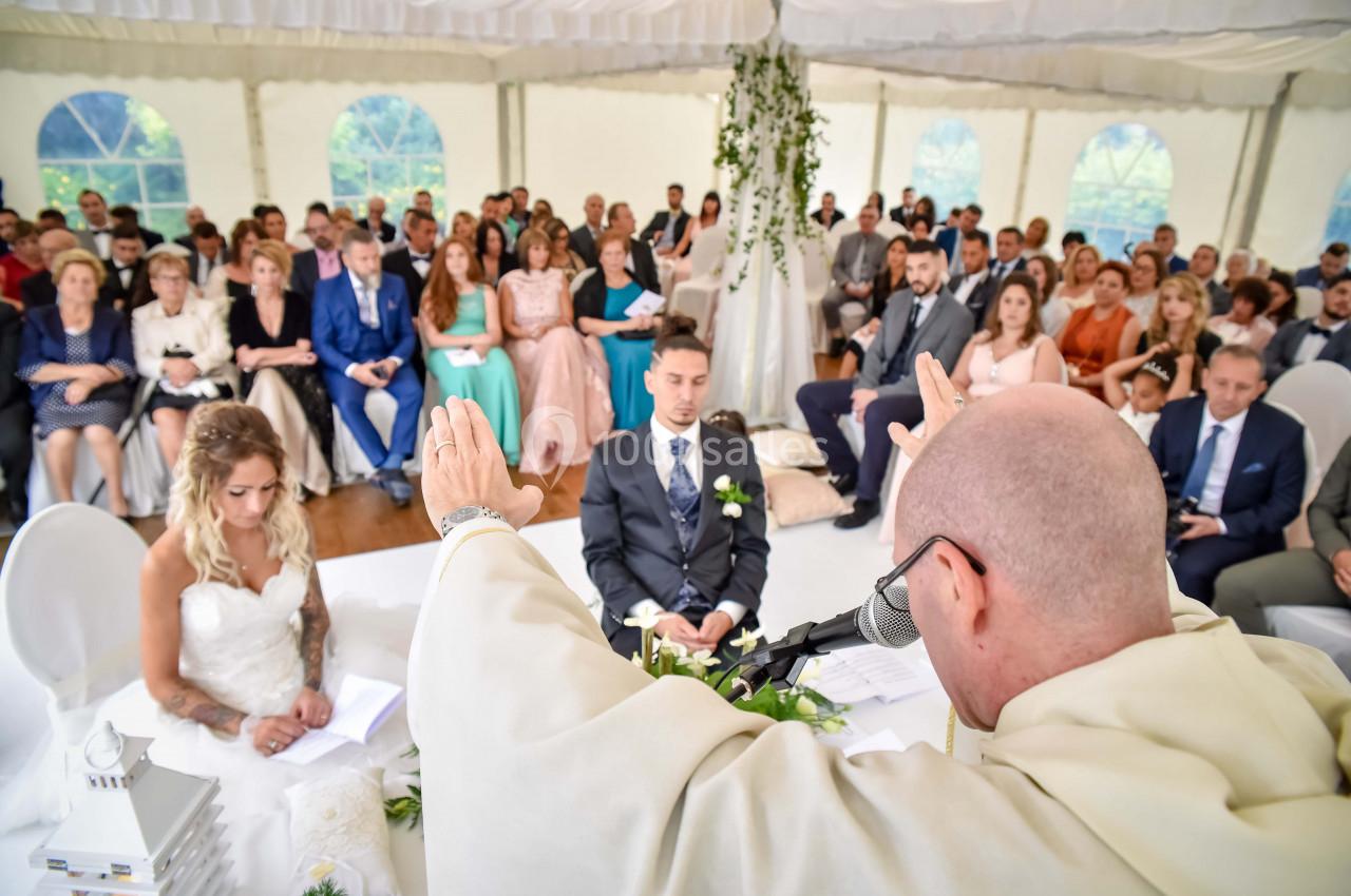 Un prêtre officie une cérémonie de mariage dans une tente, devant un couple assis et des invités attentifs.