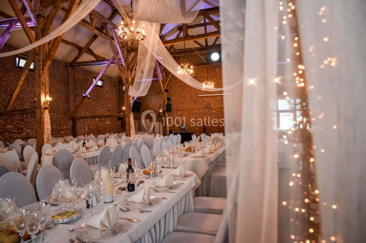 Salle de réception décorée avec des tables dressées, des chaises blanches et des guirlandes lumineuses sous une charpente en…