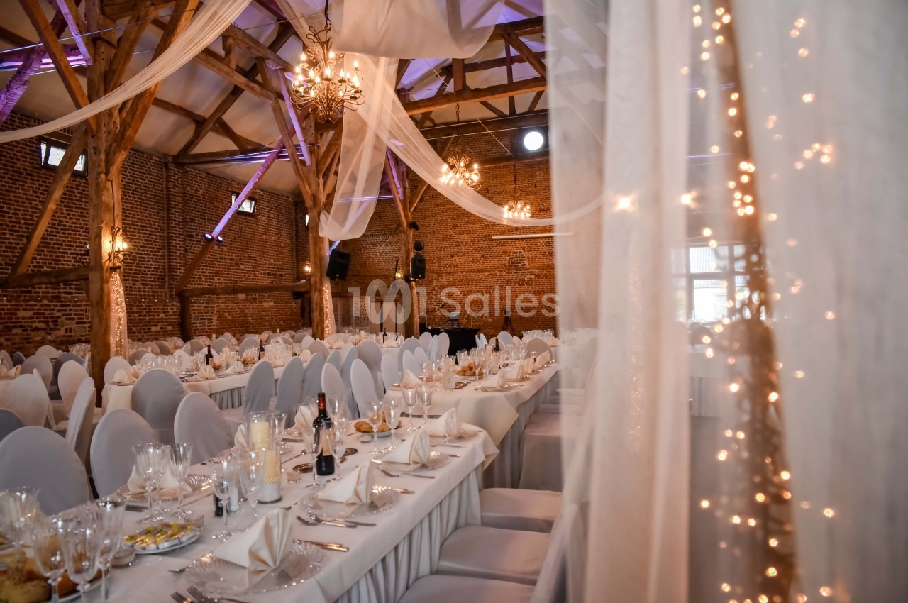 Salle de réception décorée avec des tables dressées, des chaises blanches et des guirlandes lumineuses sous une charpente en…