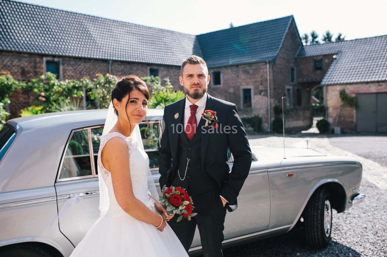 Un couple en tenue de mariage pose devant une voiture ancienne, avec des bâtiments en briques en arrière-plan.
