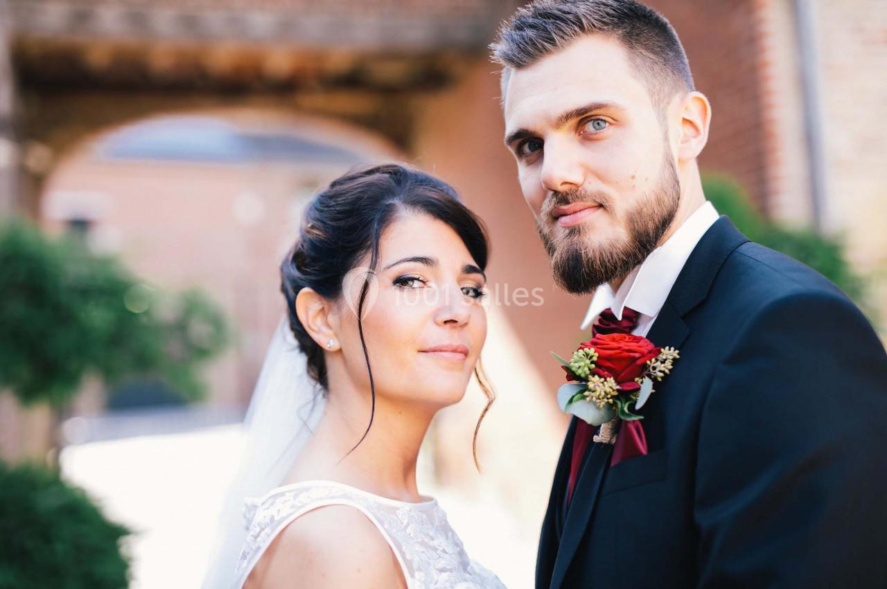 Un couple en tenue de mariage pose devant un bâtiment en briques avec des plantes en arrière-plan.