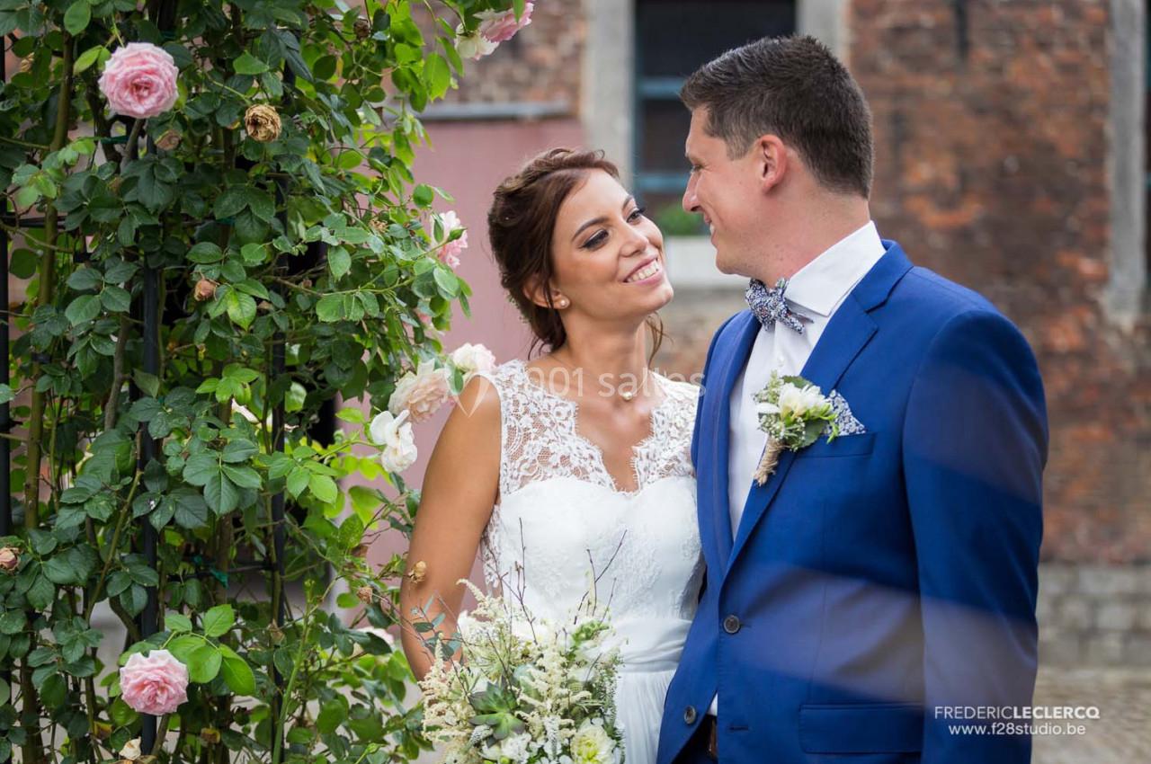 Un couple en tenue de mariage sourit près d'une arche fleurie, devant un mur en briques.