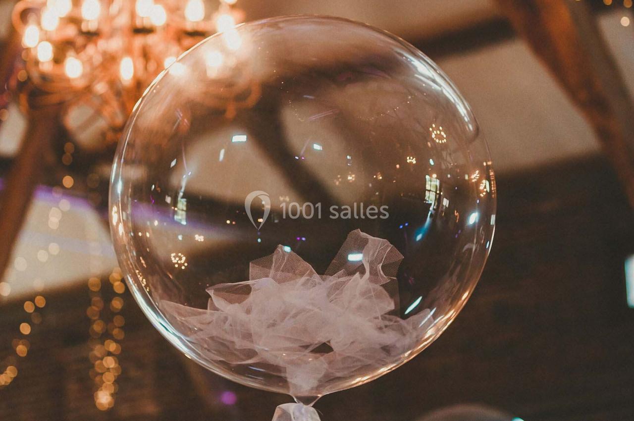 Ballon transparent décoratif avec un nœud en tulle blanc, suspendu sous un lustre dans une salle éclairée.