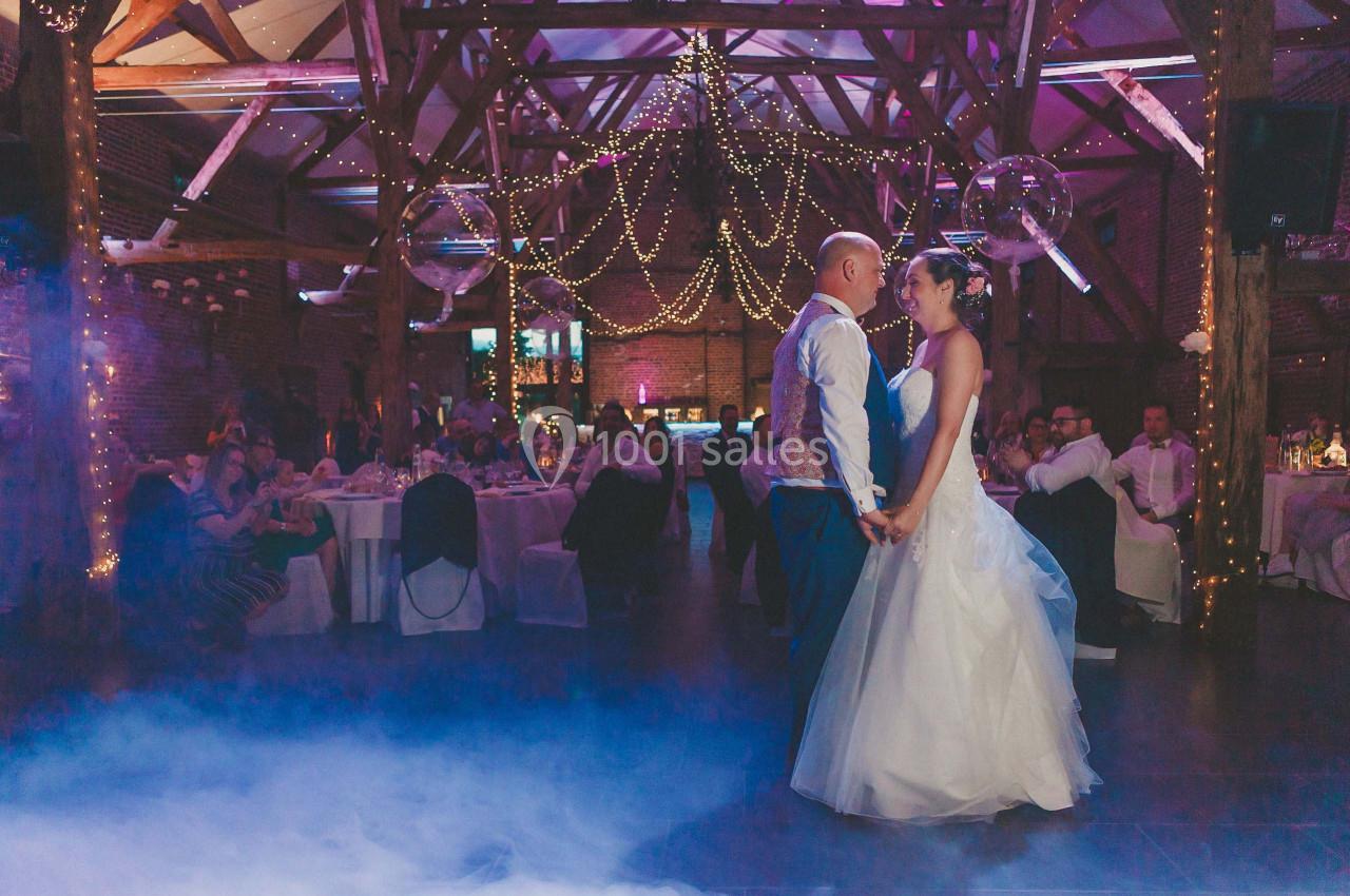 Un couple danse sous des guirlandes lumineuses dans une salle de réception en bois, entouré d'invités assis.