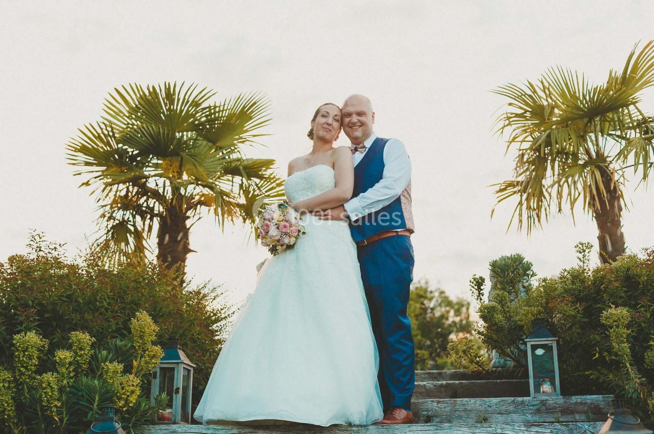 Un couple en tenue de mariage pose dehors, entouré de palmiers et de lanternes sur un escalier en bois.