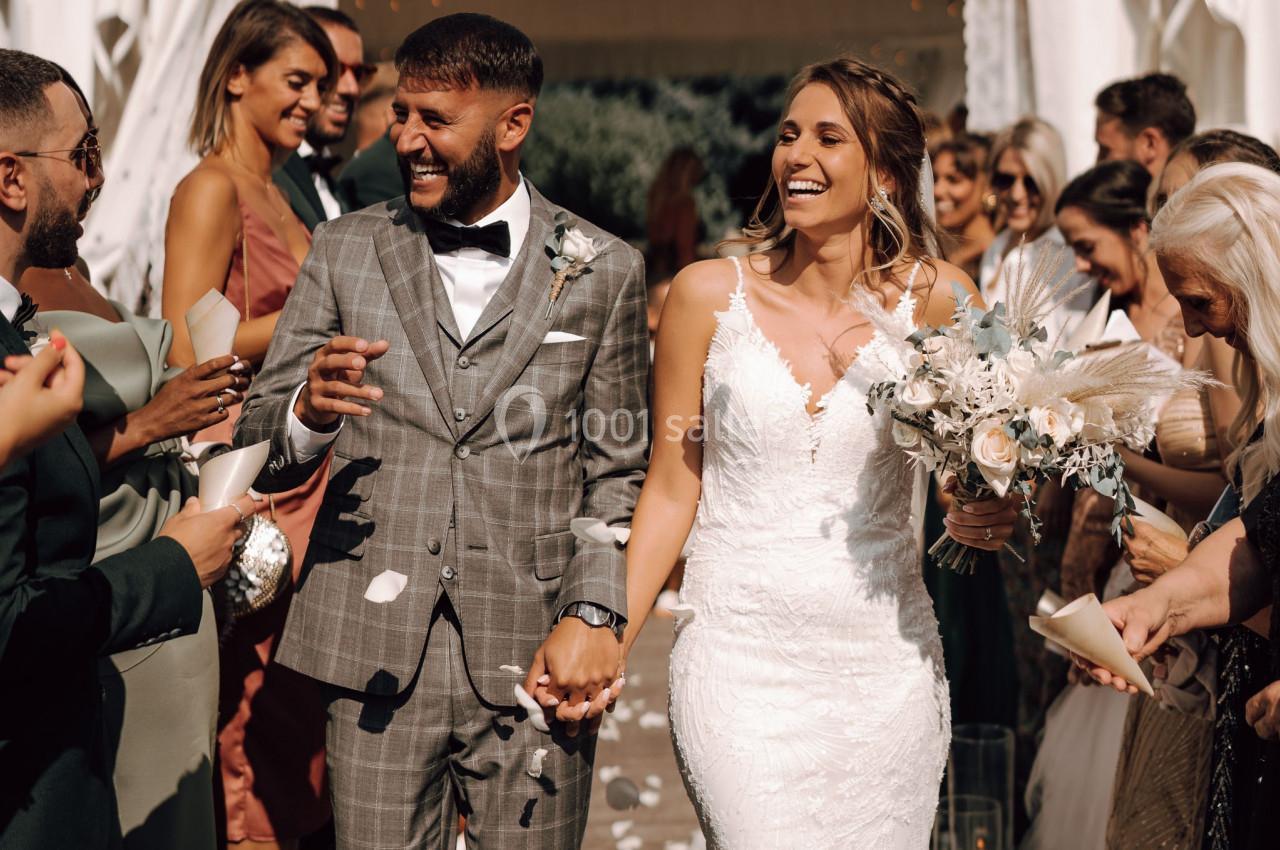 Un couple souriant en tenue de mariage marche main dans la main entouré d'invités célébrant leur union.