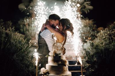 Un couple en tenue de mariage se tient la main dans un jardin baigné par une lumière douce au coucher du soleil.
