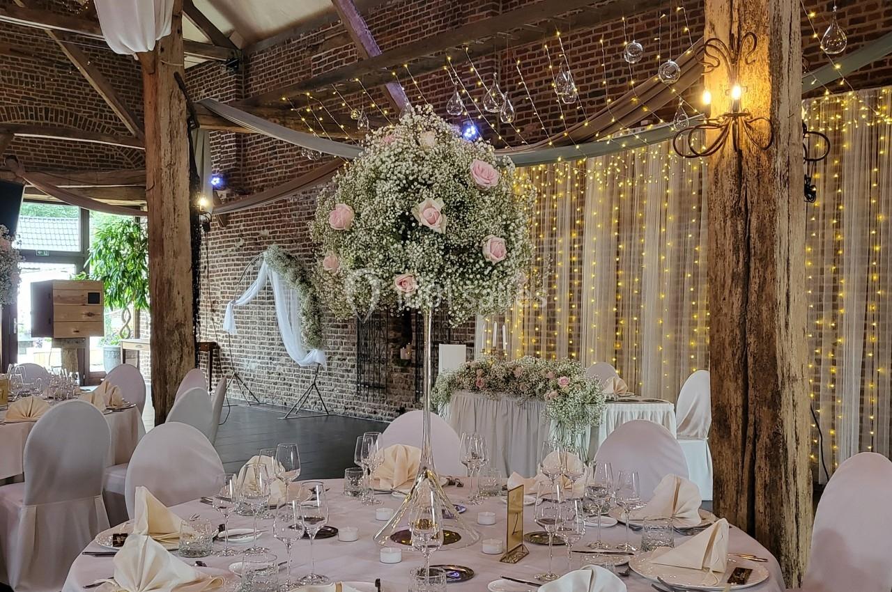Salle de réception décorée avec des tables dressées, un centre de table floral et des guirlandes lumineuses en arrière-plan.