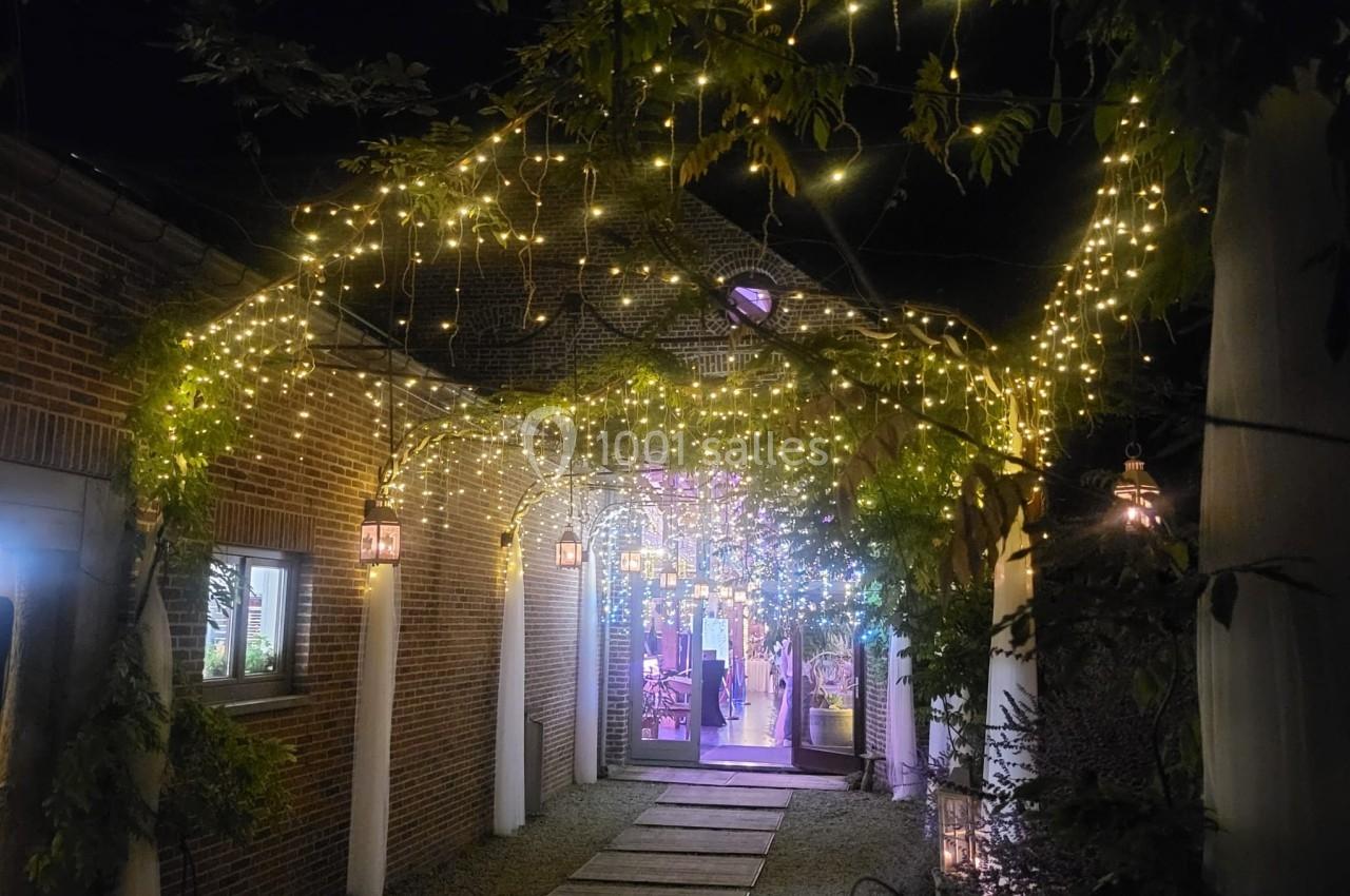 Entrée éclairée par des guirlandes lumineuses suspendues, entourée de végétation et de murs en briques.