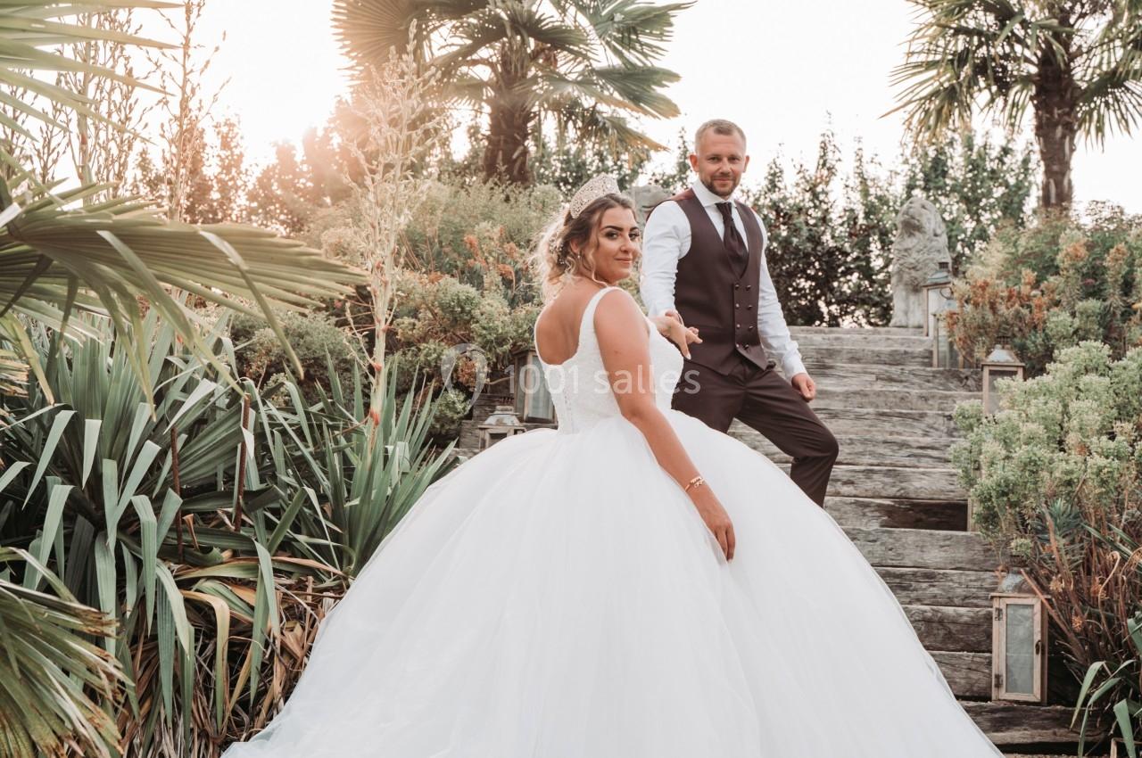 Un couple en tenue de mariage pose dans un jardin verdoyant avec des palmiers et des escaliers en pierre.