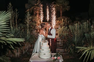 Un couple en tenue de mariage se tient la main dans un jardin baigné par une lumière douce au coucher du soleil.