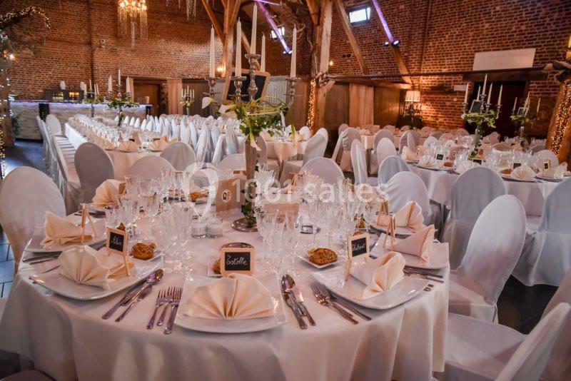 Salle de réception décorée avec des tables rondes élégantes, nappes blanches, verres et couverts soigneusement disposés.