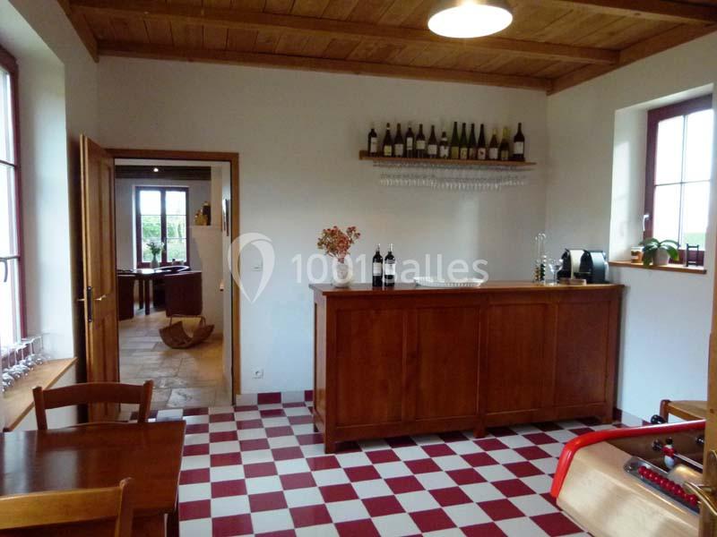 Salle avec un bar en bois, des bouteilles de vin exposées, un sol à carreaux rouges et blancs, et des fenêtres lumineuses.