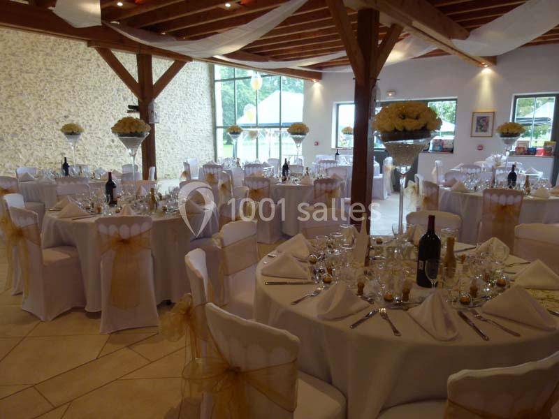 Salle de réception décorée avec des tables rondes dressées, nappes blanches et chaises ornées de nœuds dorés.