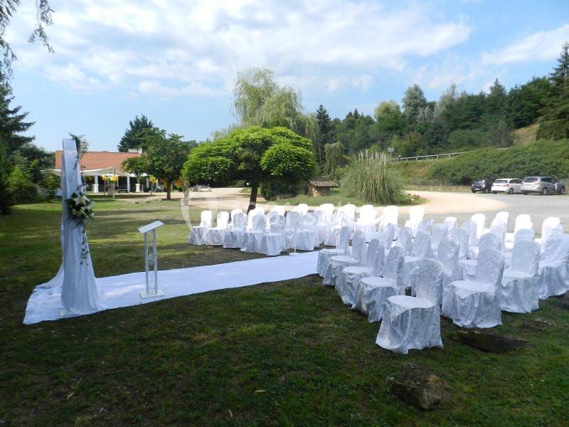 Chaises blanches alignées en extérieur pour une cérémonie, avec un pupitre et une arche décorée de fleurs.