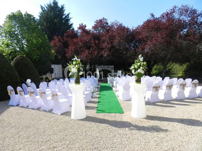 Chaises blanches alignées autour d'une allée verte, décorées pour une cérémonie en plein air devant une arche fleurie.