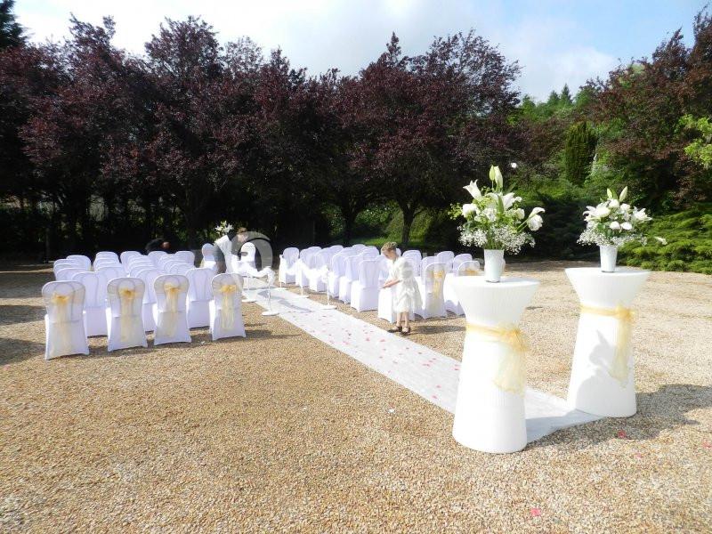 Allée centrale décorée pour une cérémonie de mariage en extérieur, avec chaises blanches et arrangements floraux.